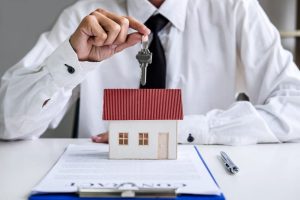 Un homme en chemise et cravate tient des clés au-dessus d'une maquette de maison posée sur un contrat de création d'entreprise