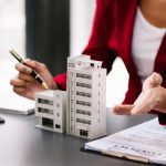 Une professionnelle en veste rouge présente une maquette d'immeuble et un contrat sur son bureau pour résoudre des litiges immobiliers