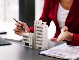 Une professionnelle en veste rouge présente une maquette d'immeuble et un contrat sur son bureau pour résoudre des litiges immobiliers