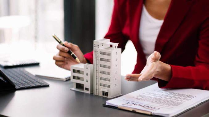 Une professionnelle en veste rouge présente une maquette d'immeuble et un contrat sur son bureau pour résoudre des litiges immobiliers